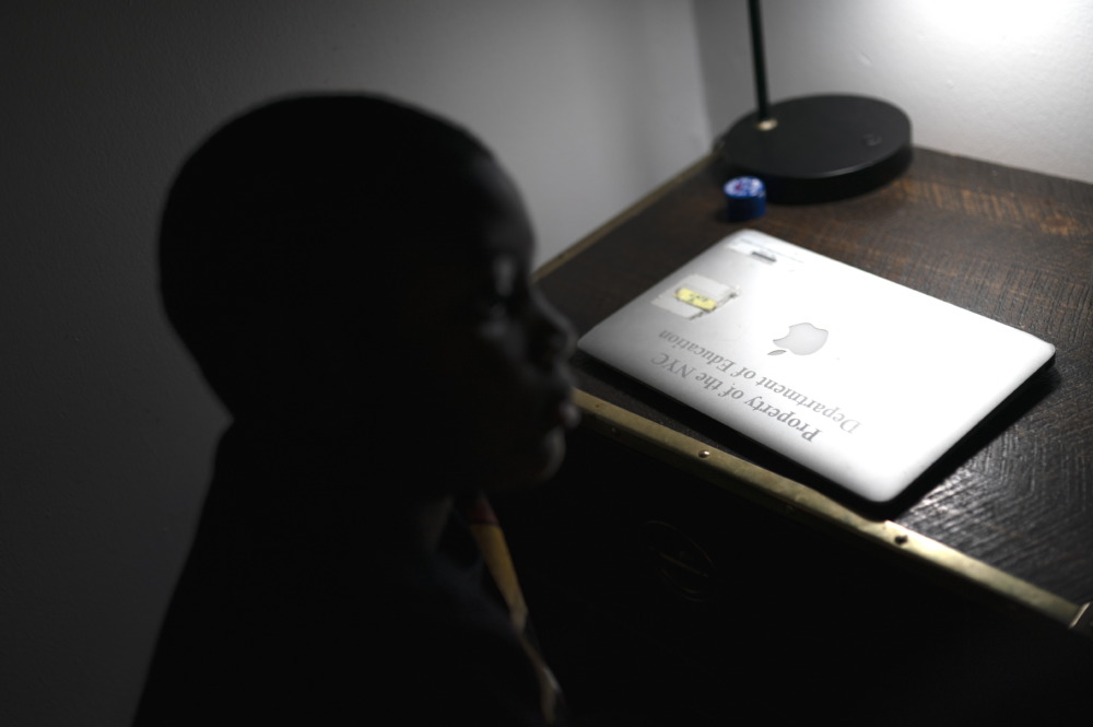 Digital divide virus: Young Black boy sits in shadows at dek with tablet showing screen reading it is property of public school district
