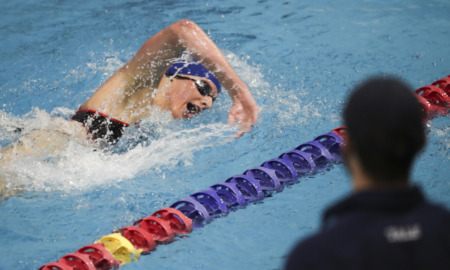 transgender athletes: White adult in bathing suit, swim cap and goggles swims in a pool next to multicolored lane divider.