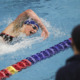 transgender athletes: White adult in bathing suit, swim cap and goggles swims in a pool next to multicolored lane divider.