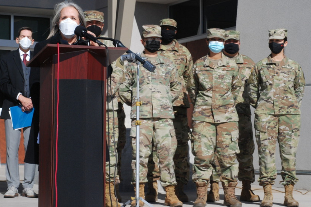 teacher shortage: Woman in red coat wearing mask stands at wood podiuom in front of 6 adult men and women in fatigue military uniforms and caps, all wearing masks