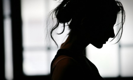 Vet Suicide Women: Woman with hair in low bun sits in profile silhouette in front of large, curtained window.
