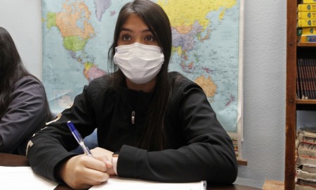 new approach to teaching race divides New Mexico: young Hispanic female student with mask on taking notes with world map on wall behind her