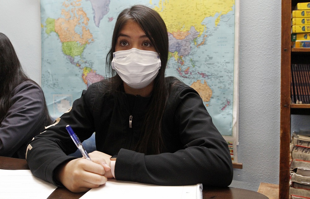 new approach to teaching race divides New Mexico: young Hispanic female student with mask on taking notes with world map on wall behind her