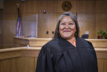New Mexico’s first Native family court: Judge Catherine Begaye, middle-aged woman with long grey hair wearing black judge's robes stands in light wood paneled courtroom smiling into camera