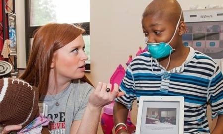 childhood cancer research grants: young red-haired woman talks to young, black boy with facemask on