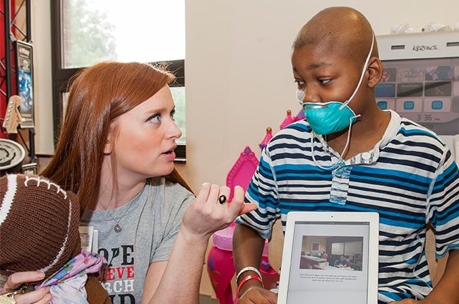 childhood cancer research grants: young red-haired woman talks to young, black boy with facemask on