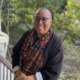 Karen Pittman opinion: Black woman with dark-frame glasses an hair pulled back stands leaning against wood porch railing wearing dark jacket and colorful scarf with greenery in the background.