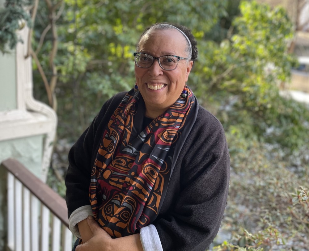 Karen Pittman opinion: Black woman with dark-frame glasses an hair pulled back stands leaning against wood porch railing wearing dark jacket and colorful scarf with greenery in the background.