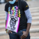 Tamir Rice: Young Black girls in black t-shirt and jeans with paper sign on chest with sketch of Tamir Rice and Black Lives Matter text stands with bows head on cement steps.