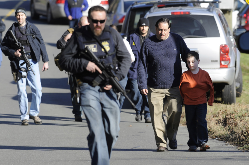 Sandy Hook Settlement: Two men wearing sunglasses and navy uniforms carrying rifles leads adults and children surrounded by parked cars walking in an asphalt parking lot.