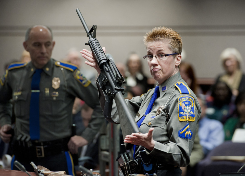 Sandy Hook Settlement: Detective Barbara J. Mattson, adult with short blonde hair wearing glasses in olive green uniform stands in crowd of people holding AR-15 rifle upright for demonstration.