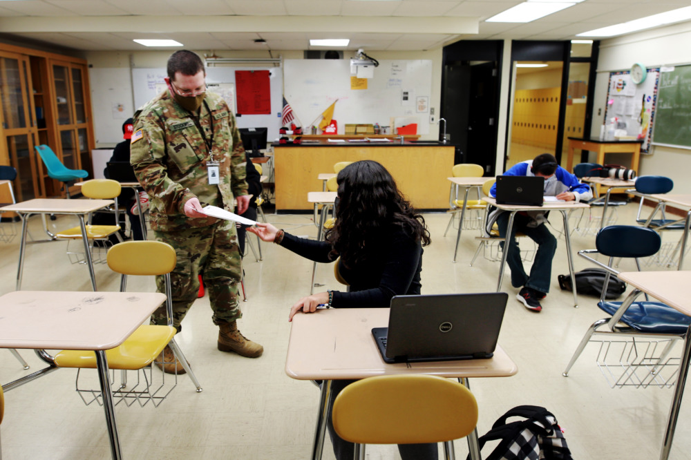 Military substitute teachers: Man in military fatigues uniform and dark mask stands in classroom handing out papers to a student sitting at desk