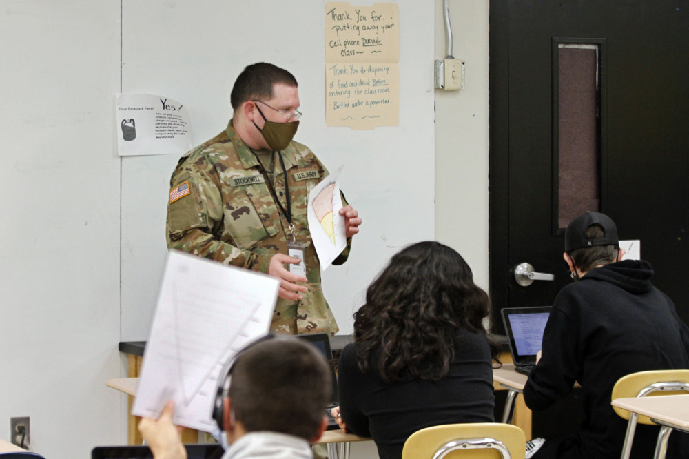 Military substitute teachers: Man in military fatigues uniform and dark mask holding white paper with red and yellow diagram stands in classroom facing several students sitting at desks with backs to camera