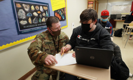 Military substitute teachers: Man in military fatigues uniform wearing black mask kneels beside male student wearing black shirt sitting at school desk in classroom