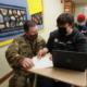 Military substitute teachers: Man in military fatigues uniform wearing black mask kneels beside male student wearing black shirt sitting at school desk in classroom