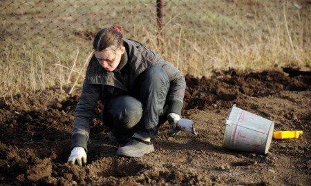 Teen facilities: Young teen crouches in garden bed wearing heavy overcoat and garden gloves prepping soil.