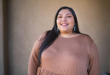 New Mexico’s Native family court: Veronica Krupnick, young woman with long dark hair weaing a taupe dress and turquoise earrings smiles into camera.