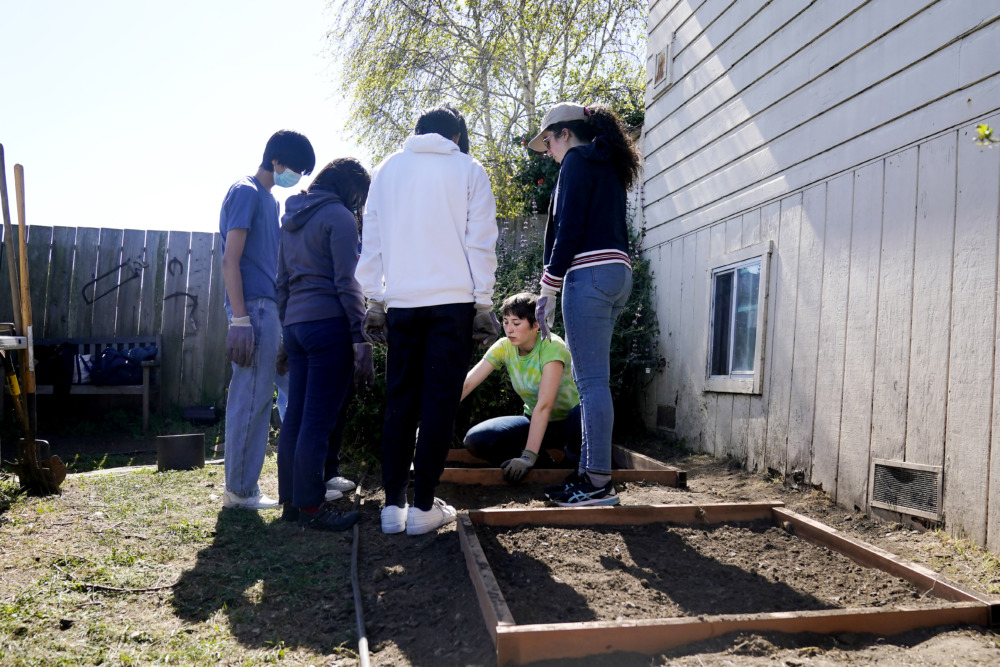student volunteers disappear: Fiv people with backs to camera stand around a crouching person who's gesturing to the soil of an unplanted garden bed with wood frame