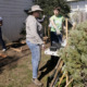 student volunteers disappear: Adult wearing jeans and light shirt and cowboy hat stands next to young woman wearing jeans and green t-shirt discussing potted plants on table in outdoor garden