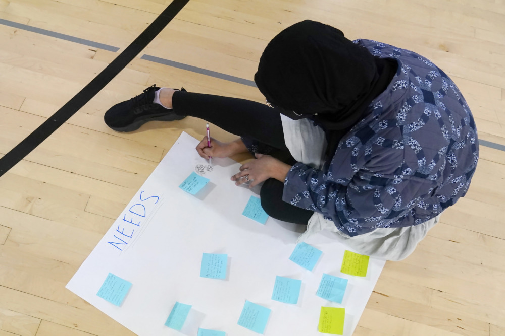 student volunteers disappear: Teen girl in dark hajib sits on light wood floor wearing dark hijab, blue print top and black leggings, writes on white poster board with multi-colored sticky notes