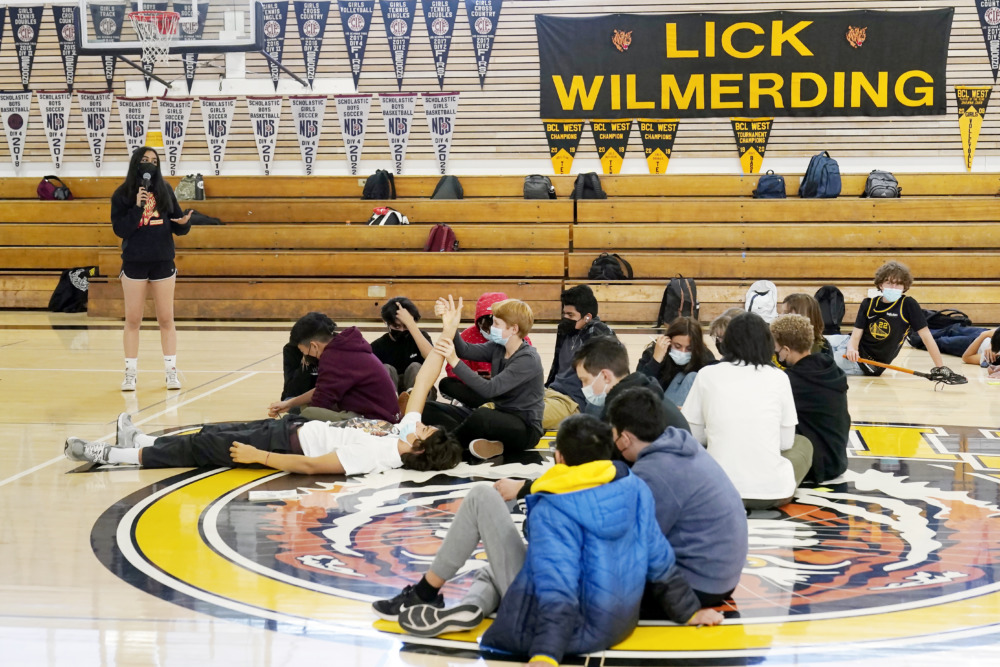 student volunteers disappear: a few dozen teens sit om wood gym floor at a meeitng