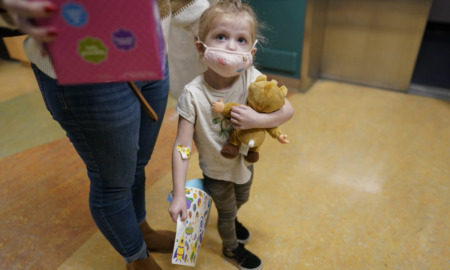 COVID and children: White, blonde toddler child wearing a pink facemask and holding a tan teddy bear stands ho.lding the hand of female adult in blue jeans and red t-shirt.