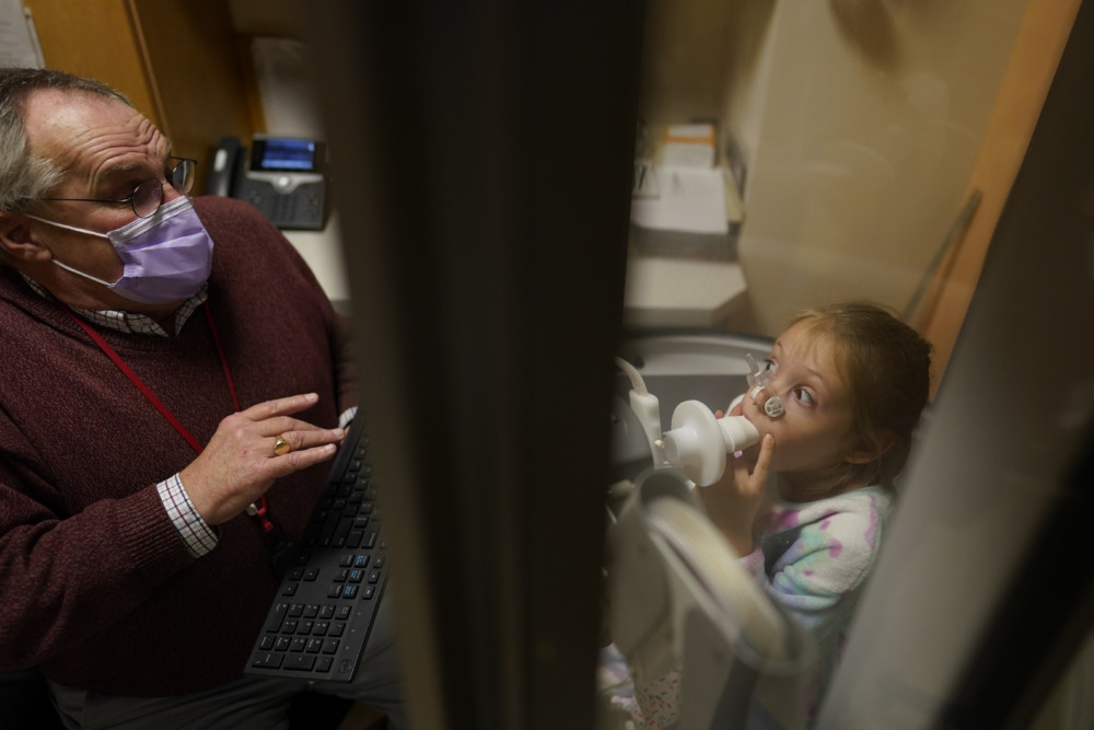 COVID and Children: Young White, blonde girl sits with large white tube held in mouth while Older male therapist in maroon lab coat administers lung capacity test.