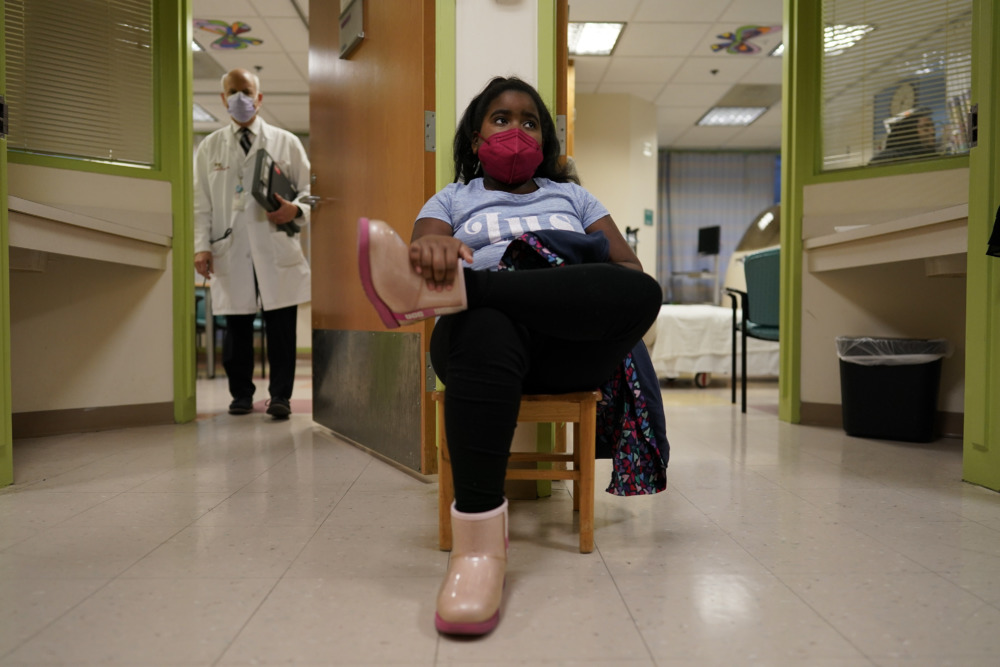 COVID and children: Young black girl with black hair wearing lavendar t-shirt, black pants, pink boots and red facemask, sits in medical office treatment room with White man in white doctor's coat wearing white facemask stands in background holding clipboards.