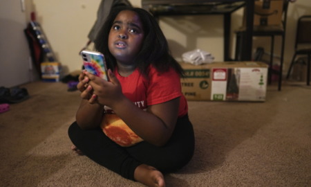 COVID and Children: Young Black girl with black hair, wearing red shirt and black pants sits on flooe holding a phone looking sad.