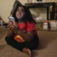 COVID and Children: Young Black girl with black hair, wearing red shirt and black pants sits on flooe holding a phone looking sad.