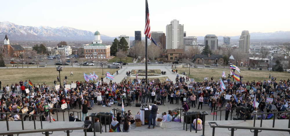 Utah Transgender Sports: Large crowd of people stand outside near an American flag with view of Salt Lake cCity skyline in the background
