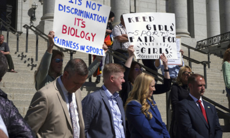Utah Transgender Sports: Several adults in business attire stand in front of adult protestors holding signs