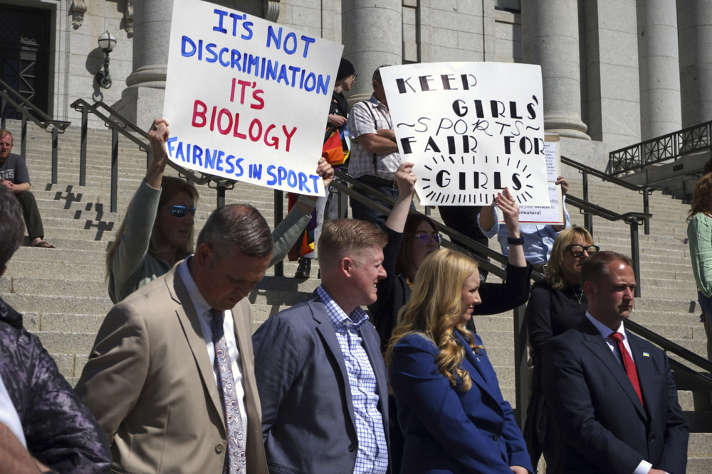 Utah Transgender Sports: Several adults in business attire stand in front of adult protestors holding signs