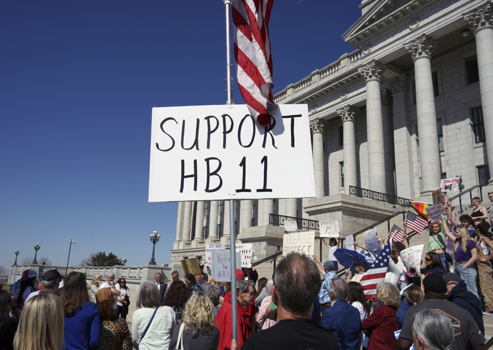 Utah Ytansgender Sports: Crowd of protesters hold sign saying, "Support HB 11"., with American flag attached to sign.