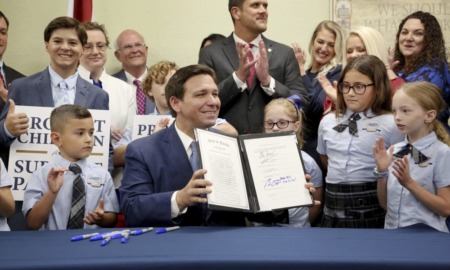 Don't Say Gay Florida: Smiling white man in blue suit sits at table holding up 2-page ocument in a display folder syrrounded by smiling white adults and children