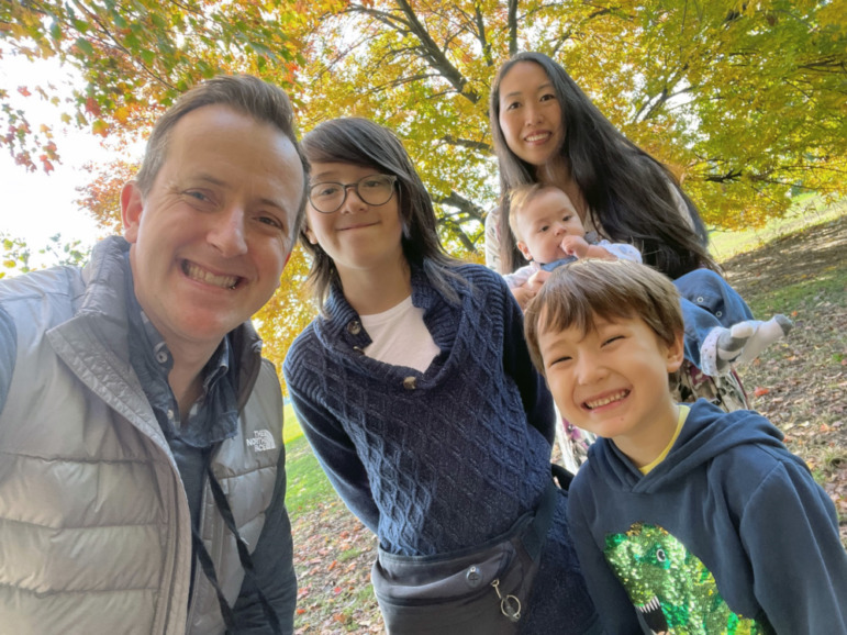 Tell kids about nuclear war: Two white adults and three white children ages infant to teenager, all with medium brown hair, stand together in front of large, leafy tree with gold foliage, all wearing sweatshirts and smiling into camera.