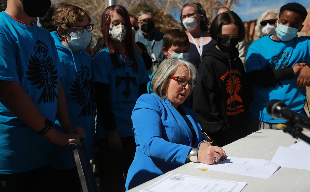 New Mexico offers equal pay to Native American teachers: older woman surrounded by people in matching blue Native logo shirts signing a document