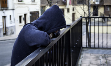 Trauma-informed therapy: Person in a dark, hooded sweatshirt leans on a black outdoor metal fence railing,