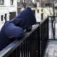 Trauma-informed therapy: Person in a dark, hooded sweatshirt leans on a black outdoor metal fence railing,