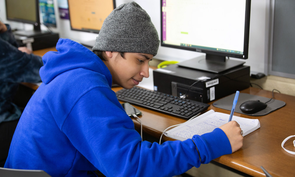 High school students cyber-defense: A young person in a blue sweatshirt and gray knit beanie writes in a notebook while he sits at a desk with a computer.