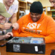 High school students cyber-defense: A young man in an orange sweatshirt sits behind an open computer case. Two young people sit on either side of him.