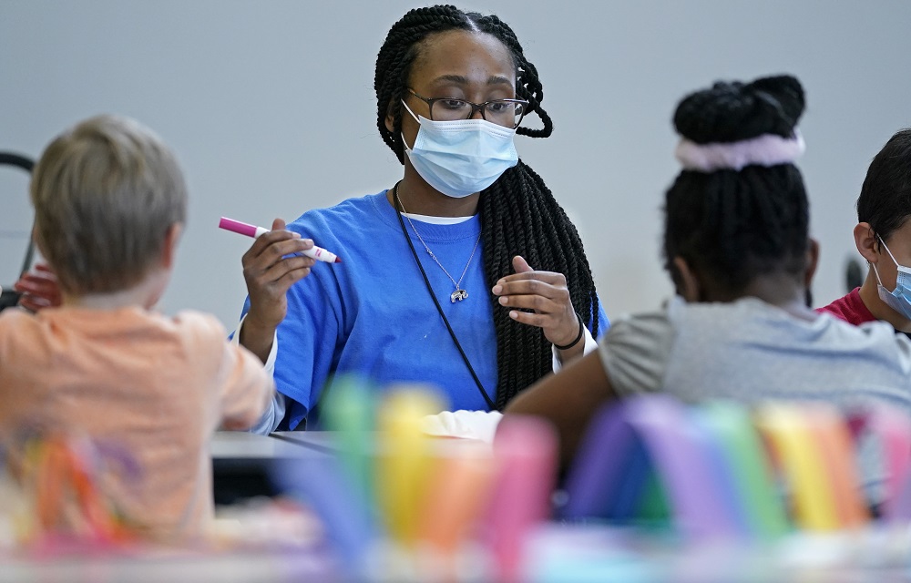 understaffing at after-school programs leaves unmet demand: black woman with long hair with facemask on works at a table with children