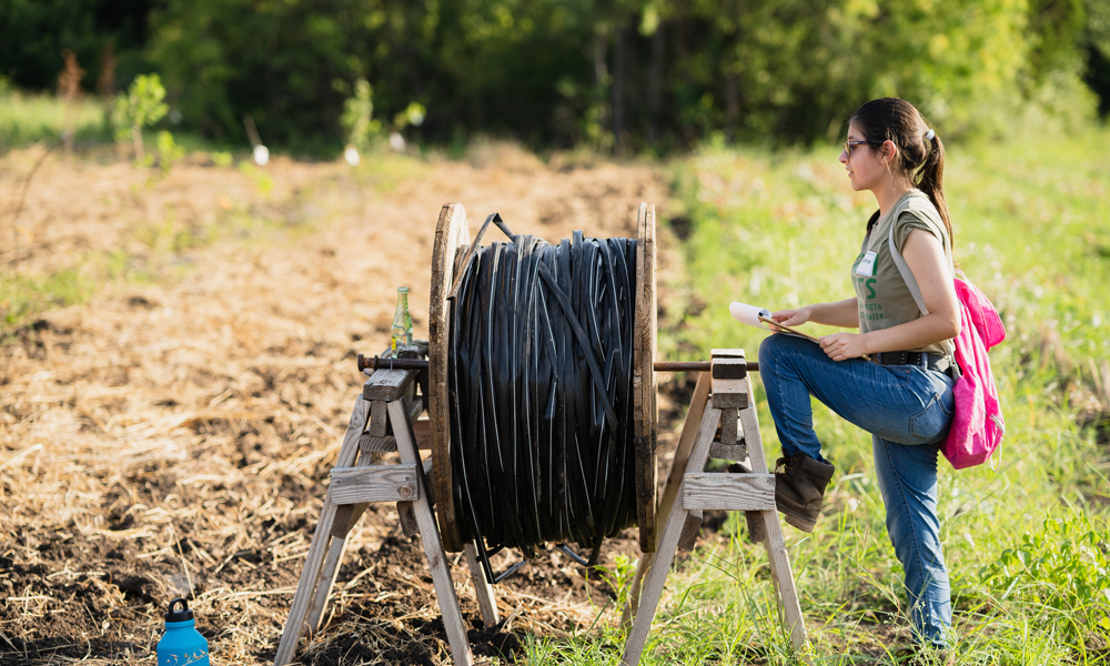 A person stands with one foot on a a wooden spool at the edge of a farming field
