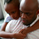 A young girl hugs her father's neck and looks over his shoulder as he, in a striped shirt, works on his computer.