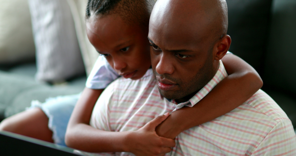 A young girl hugs her father's neck and looks over his shoulder as he, in a striped shirt, works on his computer.