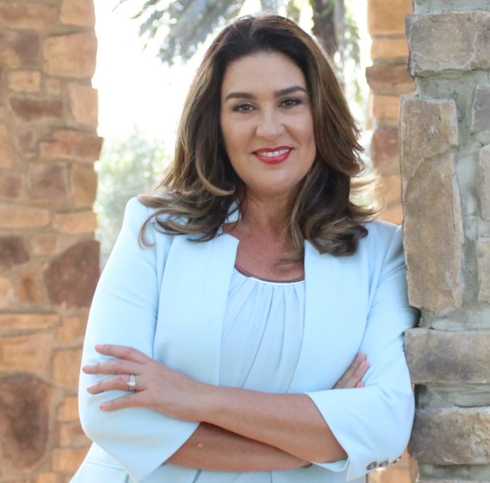 Nonprofit ladership: Dark-haired woman wearing whit jacket and blouse, stands with arms crossed between two pale tan stonework pillars