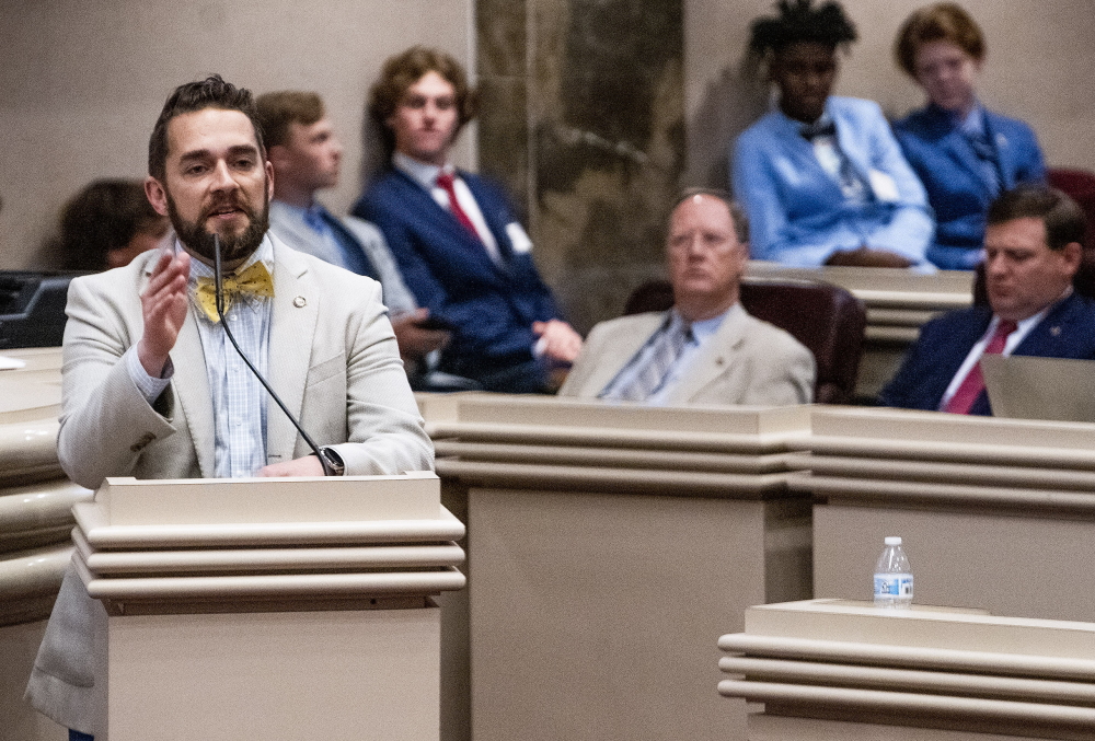 Trans teens Alabama: White man stands at podium speaking into microphone with seated adults also behind a light wood podiums surround him.