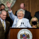 Transgender youth Alabama: Older, gray-haired woman wearing light blue suit raises right arm behind brown podium with Alabama state seal surrounded by men in dark suits