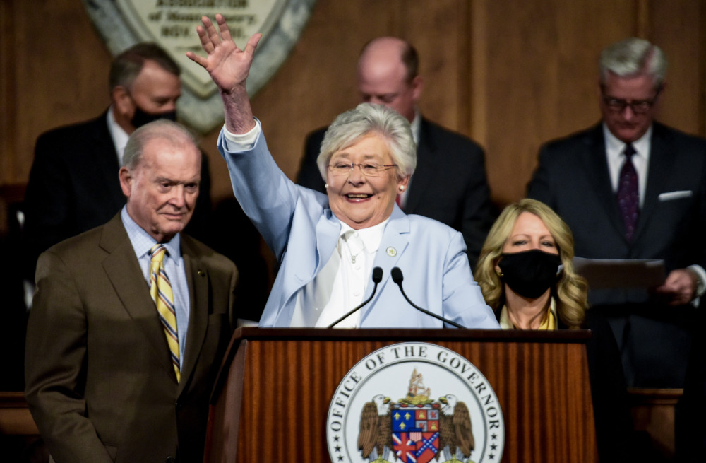 Transgender youth Alabama: Older, gray-haired woman wearing light blue suit raises right arm behind brown podium with Alabama state seal surrounded by men in dark suits