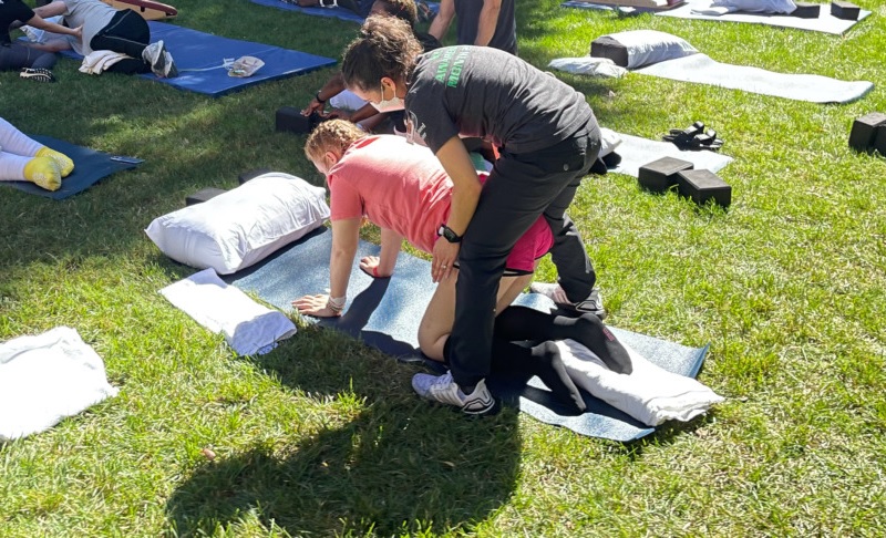 adaptive yoga: A partially paralyzed teen girl, wearing an orange shirt and shorts, gets help from an adaptive yoga instruction who is supporting and hold up the girl's torso, while the girl is on her hands and knees on a yoga mat.